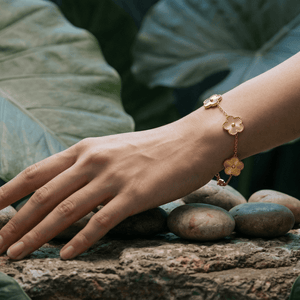 A woman's hand wearing a Flower Charm Bracelet rests on stones among lush green leaves.