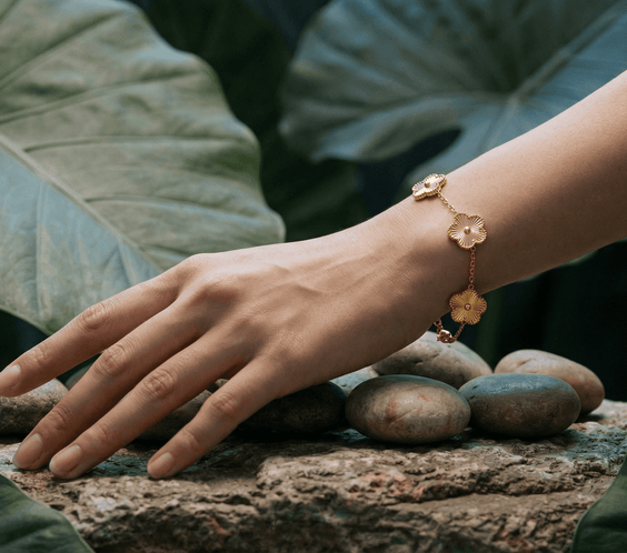 A woman's hand wearing a Flower Charm Bracelet rests on stones among lush green leaves.