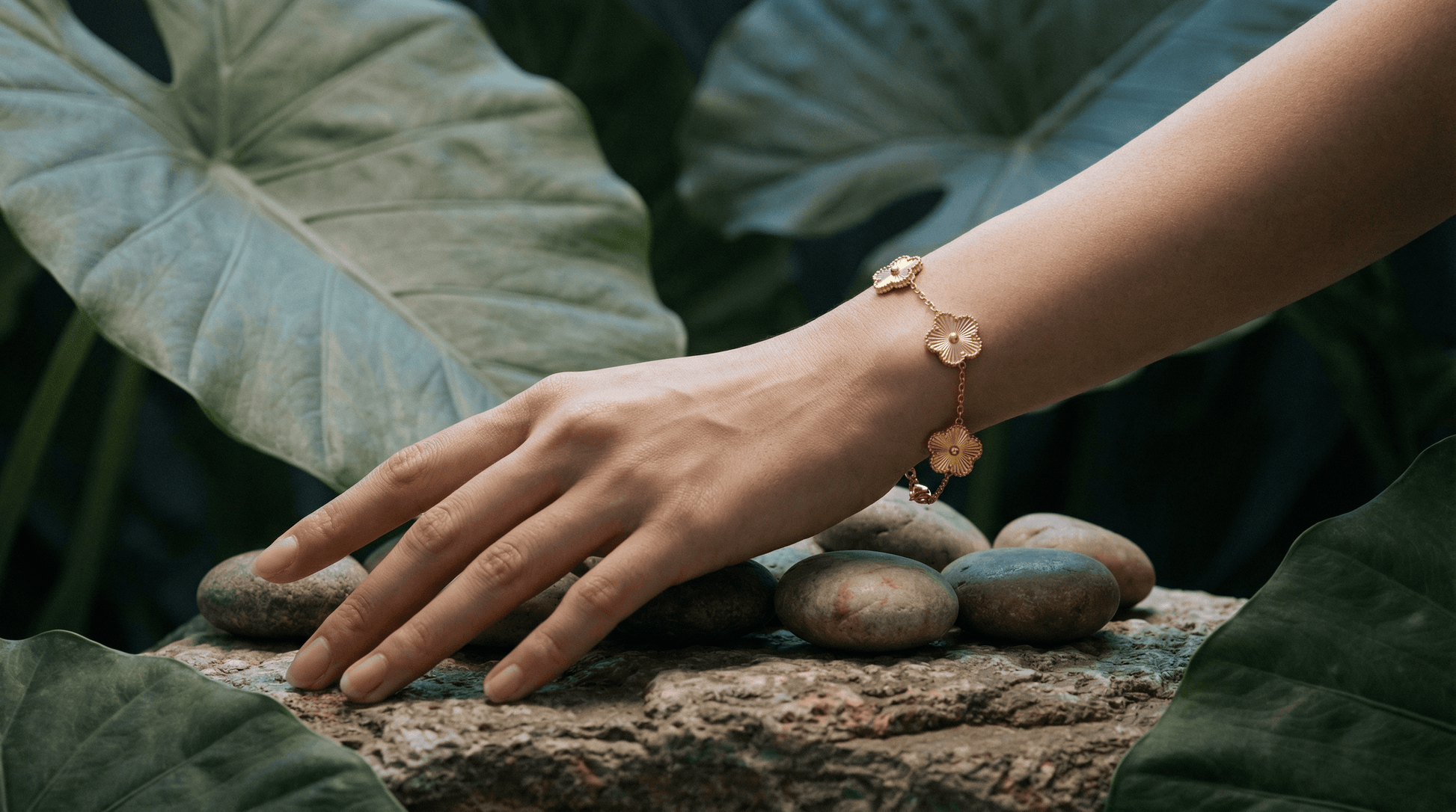 A woman's hand wearing a Flower Charm Bracelet rests on stones among lush green leaves.