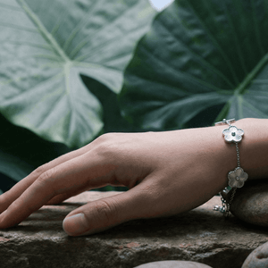 Close-up of a hand wearing a Flower Charm Bracelet on natural stones, surrounded by large green leaves.