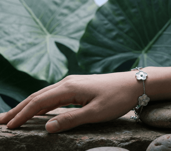 Close-up of a hand wearing a Flower Charm Bracelet on natural stones, surrounded by large green leaves.