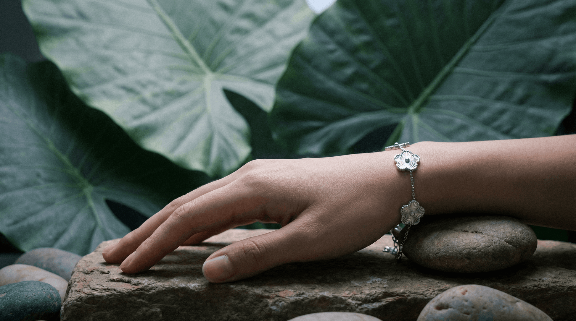 Close-up of a hand wearing a Flower Charm Bracelet on natural stones, surrounded by large green leaves.