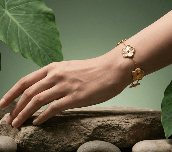 A hand wearing a Flower Charm Bracelet, surrounded by stones and green leaves, showcasing elegant floral jewelry.