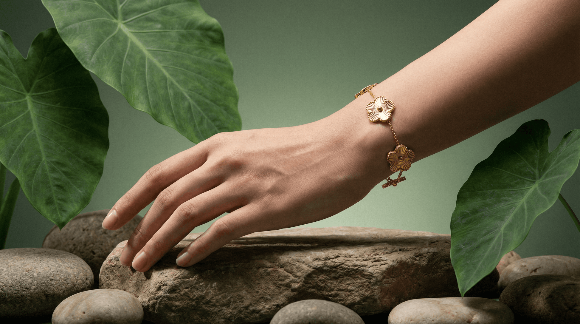 A hand wearing a Flower Charm Bracelet, surrounded by stones and green leaves, showcasing elegant floral jewelry.