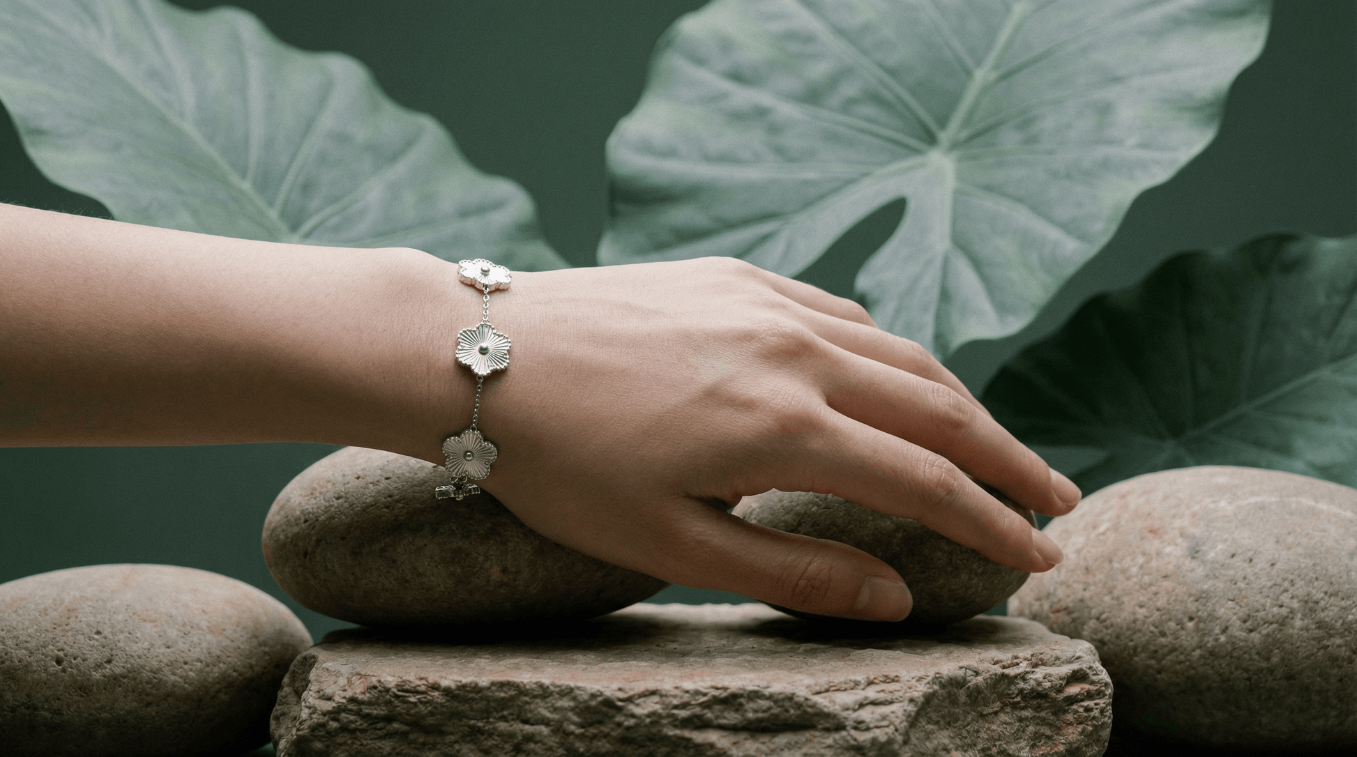 Flower Charm Bracelet worn on a hand resting on stones with green leaves in the background.